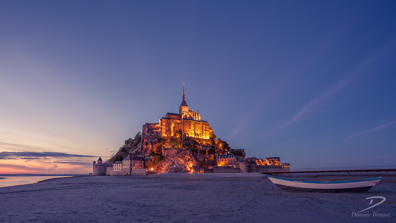 Wide-angle photo of Mont St. Michel at dusk (blue hour) with a small boat in the foreground